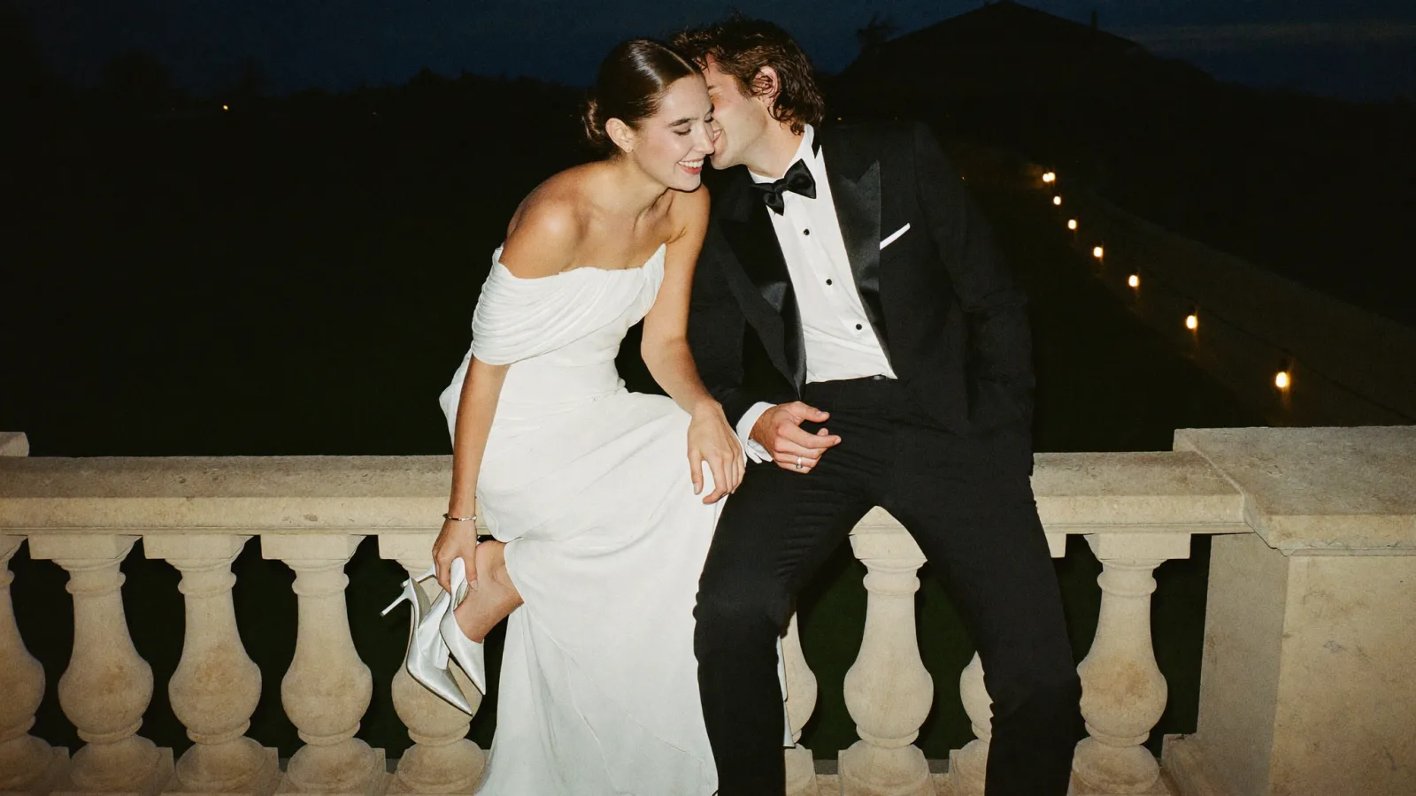 Bride and groom laughing on a stone balustrade at night, the bride holding her heels